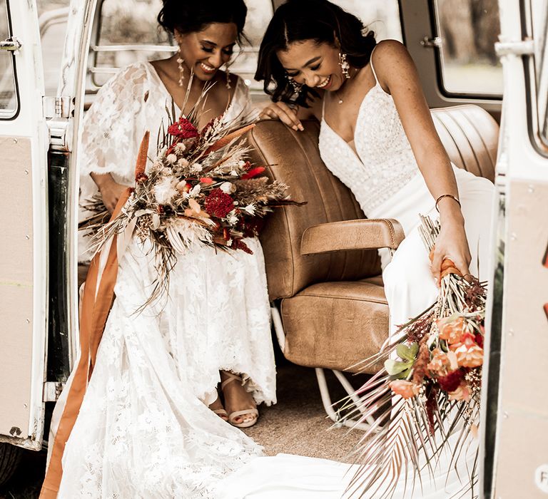 Two brides sit in a camper van and laugh at a boho wedding shoot. they both wear lace boho inspired dresses. They hold dried flower bouquets.