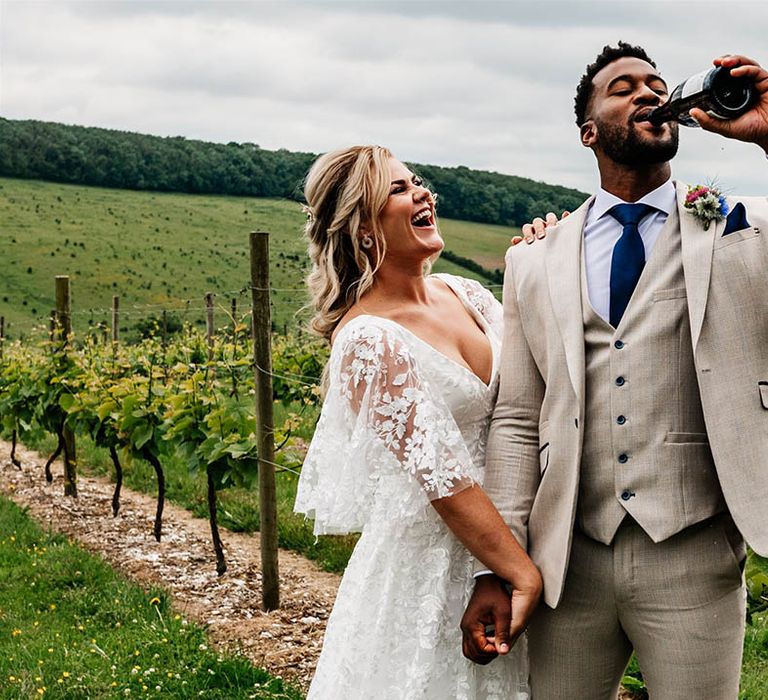 Groom in a beige three-piece suit drinking champagne with his bride in a lace wedding dress holding his arm 