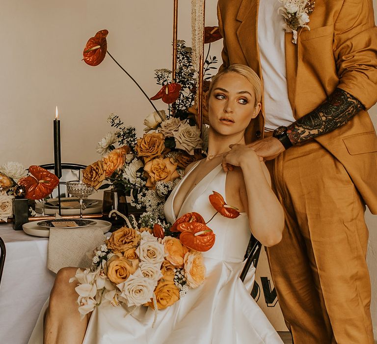 Groom in a white t-shirt and mustard suit with tattoos placing his hand on his brides shoulders at their intimate wedding reception 