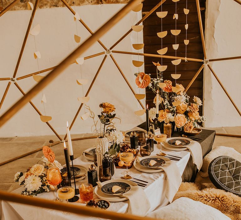 A relaxed wedding reception table with cushions and antipasti under a wooden geodome structure with hanging garlands 