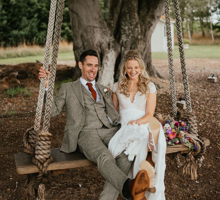 Bride in lace top cap sleeved wedding dress sits with groom in grey three piece herringbone suit and red tie on giant wooden swing outside during late summer wedding at Wellington Wood Norfolk