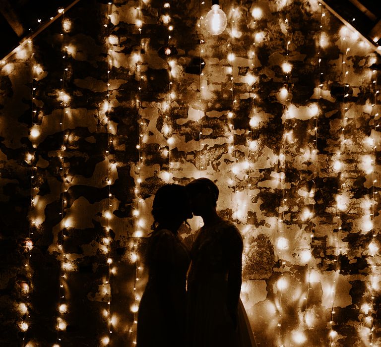 Portrait of two brides kissing in front of a curtain of fairy lights