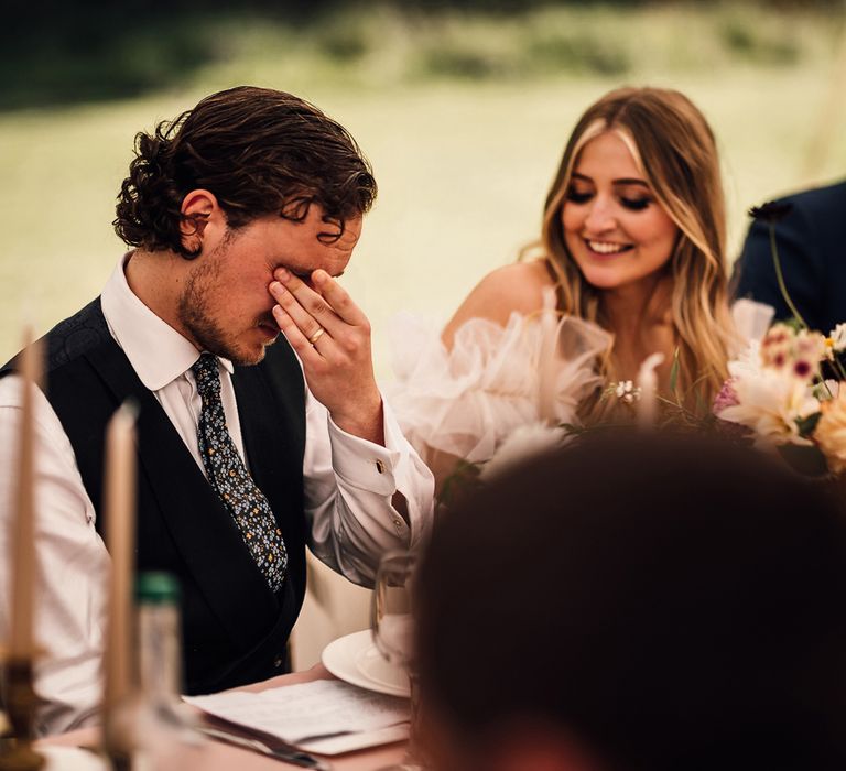 Emotional groom in white shirt, blue waistcoat and patterned tie holds hands to eyes as bride in Halfpenny London Mayfair dress smiles during marquee wedding reception in Cornwall