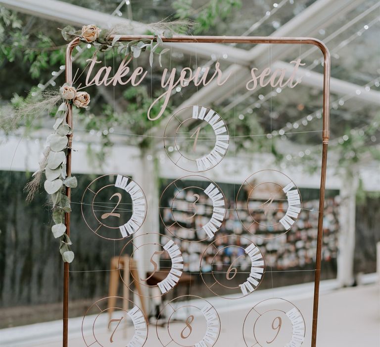 Copper frame seating plan with circular numbers and names strung across and foliage decor for marquee wedding reception