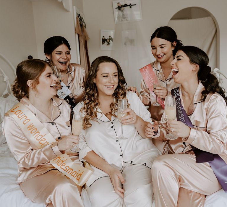 Bride in white satin pyjamas sits on double bed with bridesmaids in ivory satin pyjamas and sashes as they all hold glasses of sparkling wine before DIY garden wedding in Bedfordshire