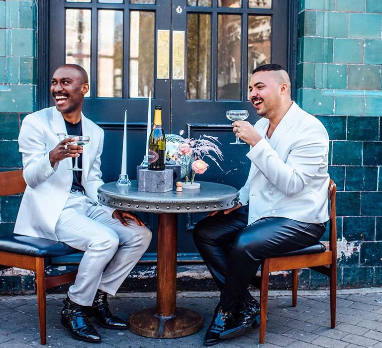 Groom in a white satin suit sipping champagne with his groom in black leather trousers and white blazer at their city elopement 