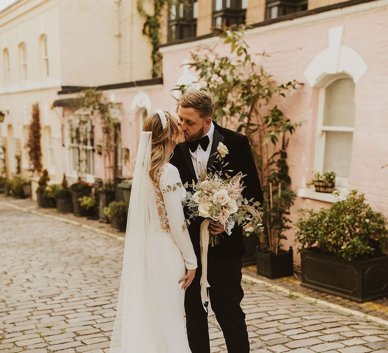 Groom in a tuxedo kissing his bride in a lace back wedding dress and cathedral length veil on the cobbled streets in London 