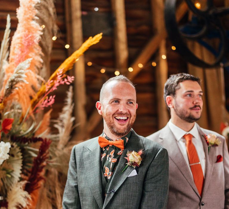 Groom in green patterned shirt, green three piece suit and orange bow tie smiles as he waits for bride to walk down the aisle at barn wedding ceremony 