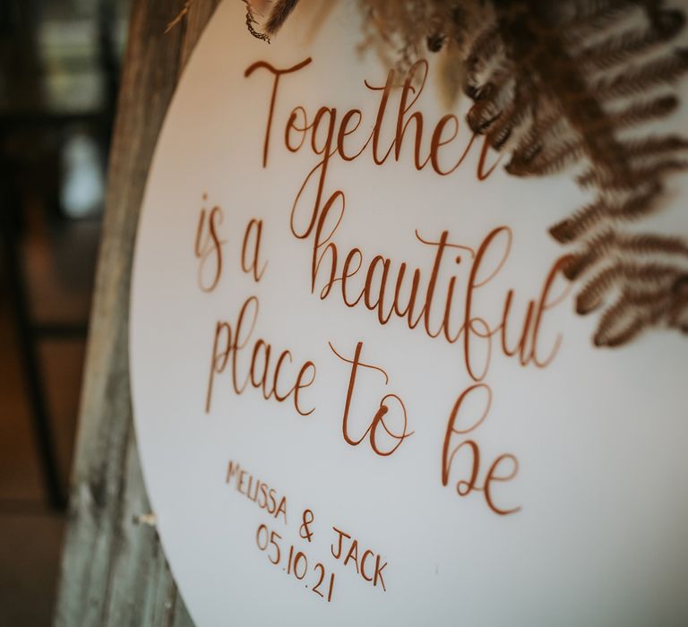 Round wedding sign reading 'Together is a beautiful place to be' on wooden board with ferns for rustic wedding