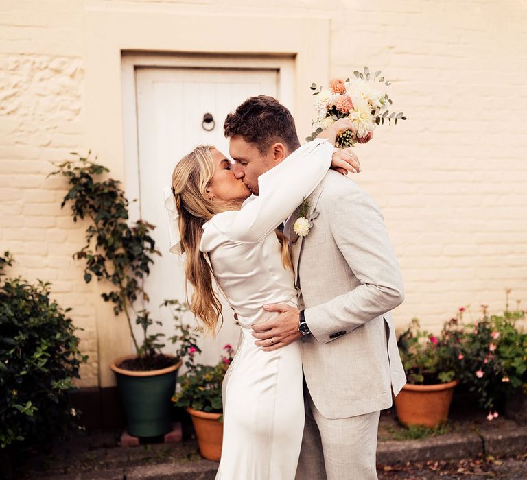 Bride in satin The Own Studio wedding dress holding white and pink bridal bouquet kisses groom in grey suit and brown suede shoes outside white painted house 