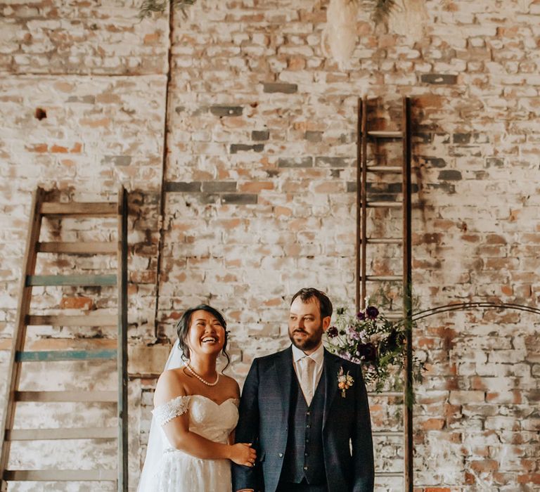 Bride in lace wedding dress and pearl necklace laughs whilst holding arm of groom in blue check three piece suit and light pink tie during wedding ceremony