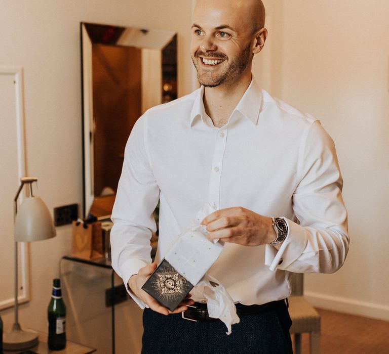 Groom in white shirt and dark trousers opens gift before wedding