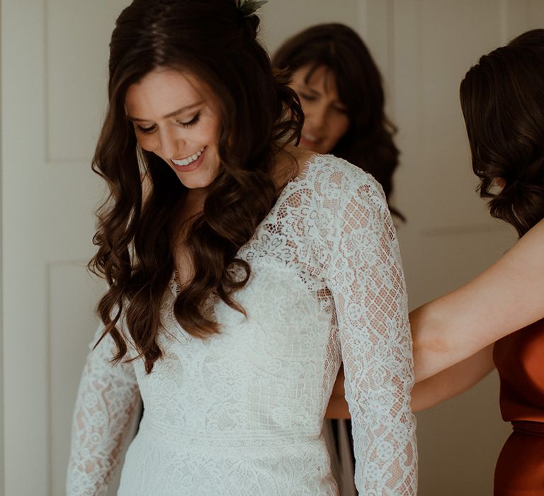 Bridesmaids in orange satin dresses help the bride into her lace wedding dress 