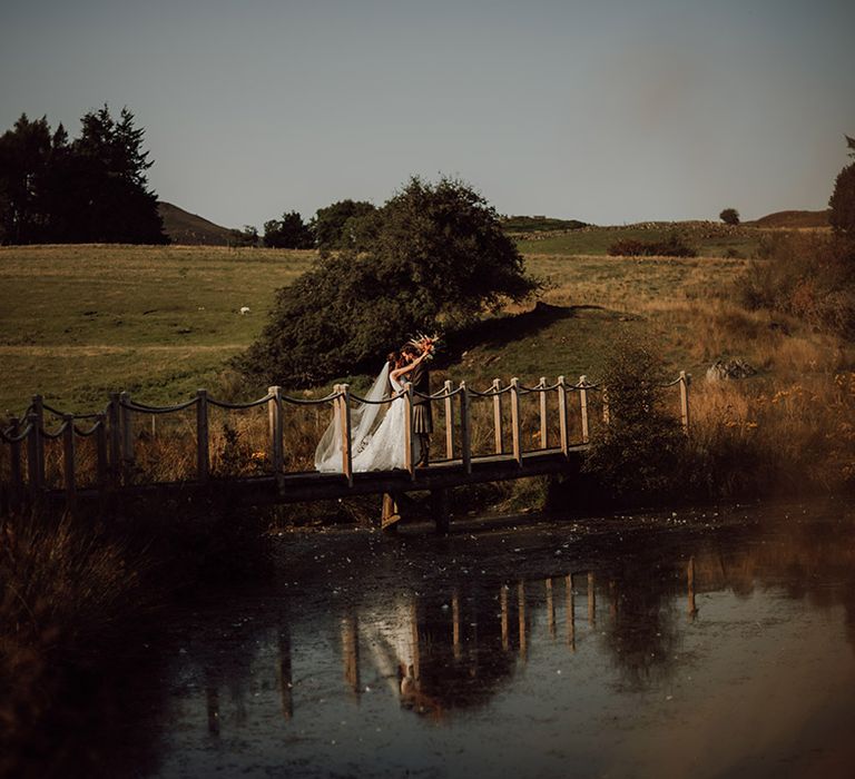 Bride in lace skirt wedding dress kisses with groom on bridge abve loch for wedding at Cardney Estate in Scotland