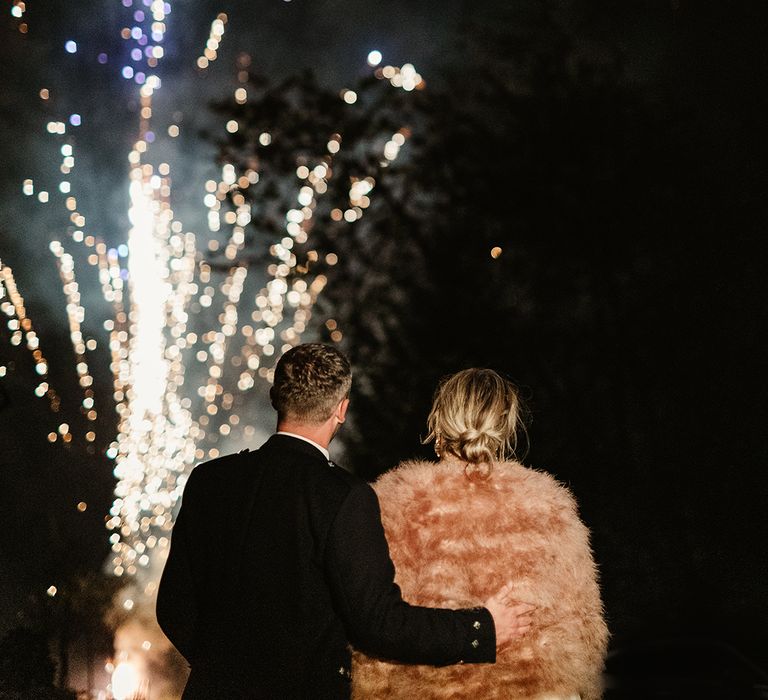 Bride in pink fur jacket and groom in black tie and kilt stands with arm around bride as they watch fireworks at the end of their wedding day