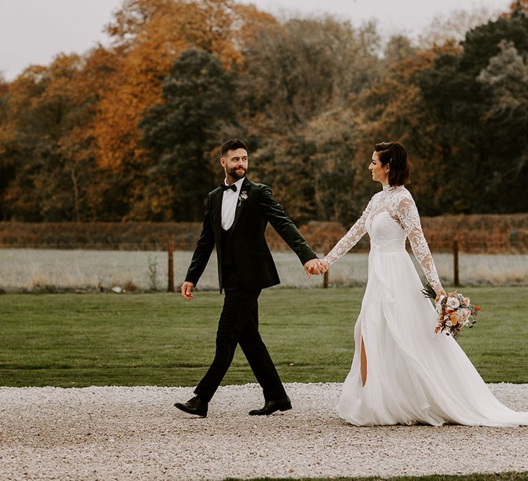 Bride and groom walk around their wedding venue together with bride in lace long sleeve wedding dress and groom in velvet black tie 