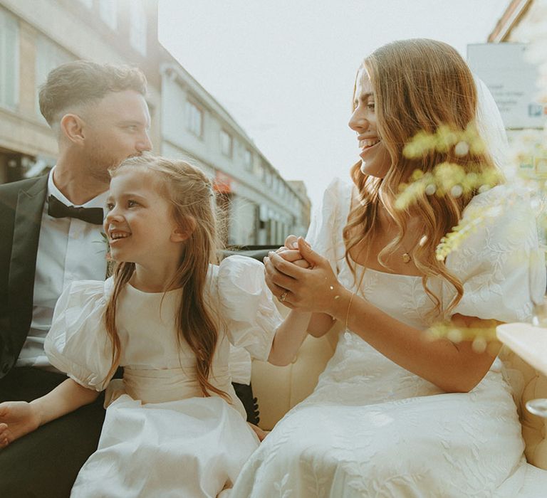 Groom in black tie sits in carriage ride over to the wedding reception with bride holding hands with their daughter, a flower girl 