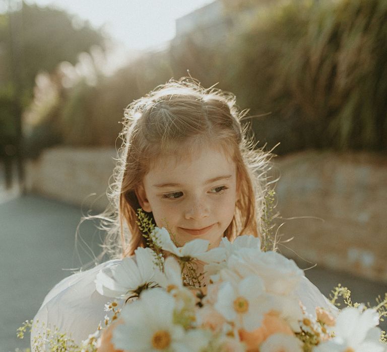 Flower girl in white dress holding the bridal bouquet with pink and white flowers