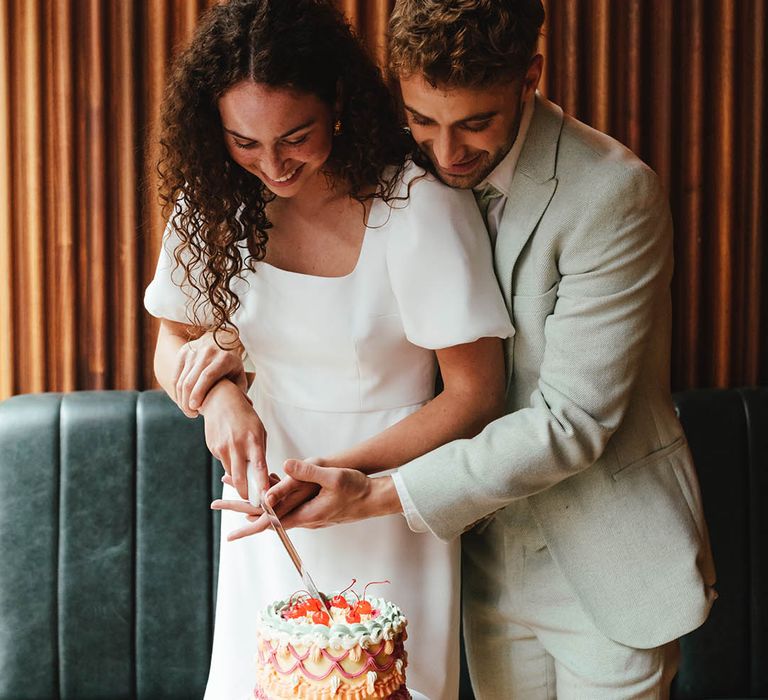 Groom in a pale green suit and bride in a short wedding dress cutting their buttercream retro wedding cake 