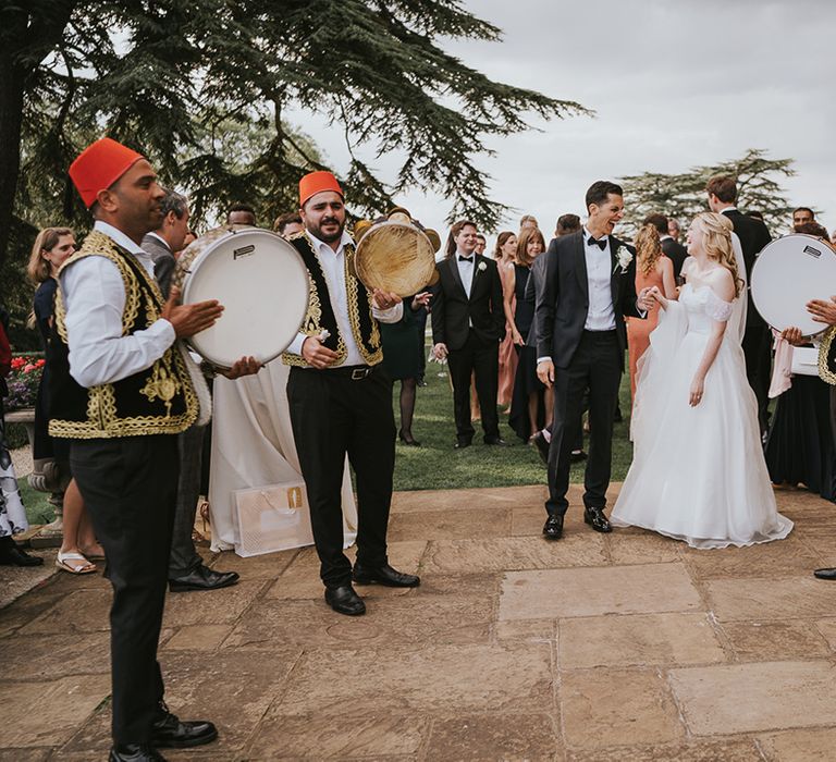 Bride and groom laugh outside their wedding venue with their Zaffa band 