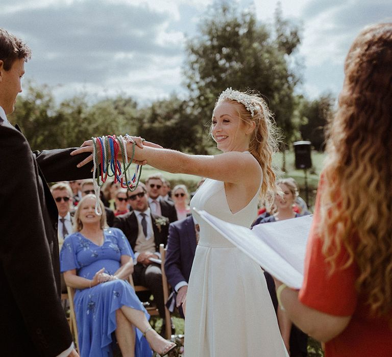 Bride and groom participate in hand fasting ceremony on their wedding day with red, green, blue and white ropes 