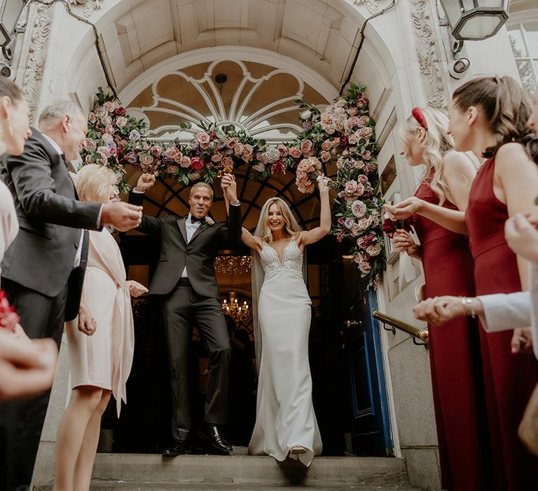 Bride and groom exit Chelsea Old Town Hall decorated with pink flowers to bridesmaids in dark red dresses 