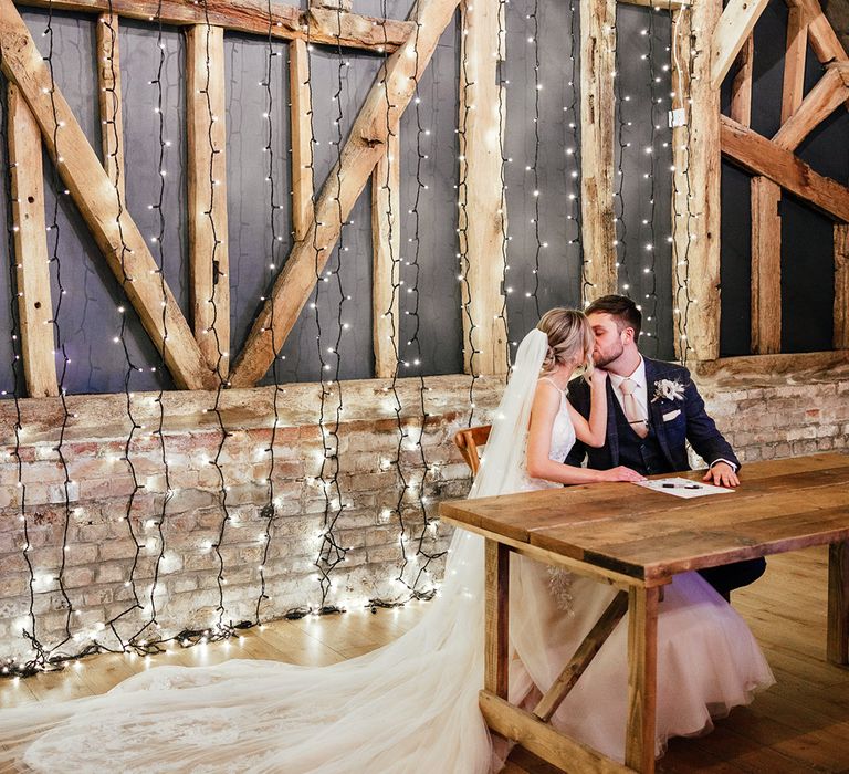 Bride and groom kiss as they sign the register in their rustic barn wedding venue decorated with fairy lights 