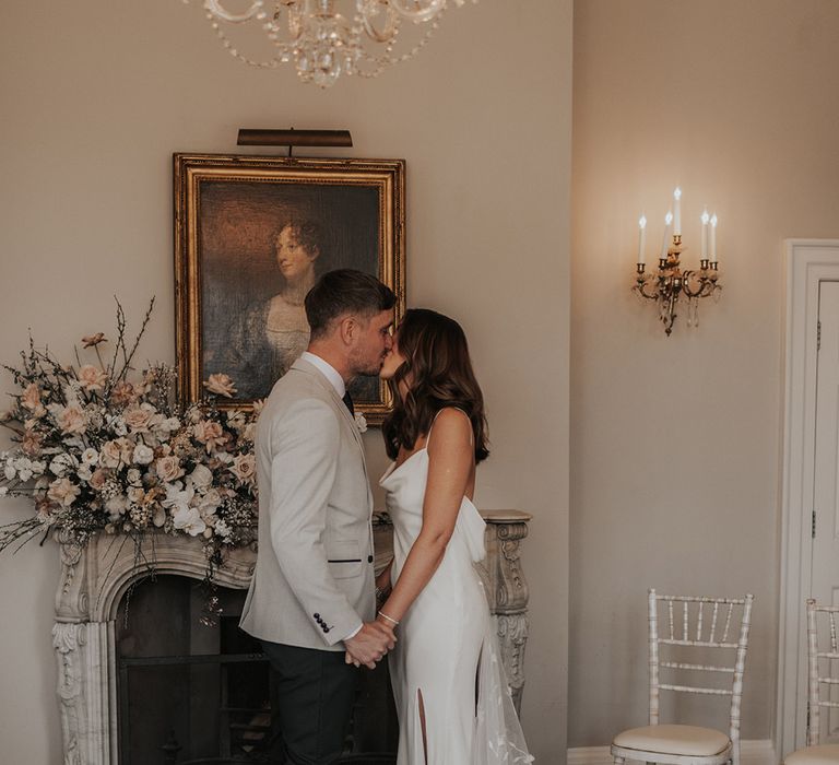 Bride and groom share a kiss in front of a pretty fireplace with white and pink wedding flower arrangement 