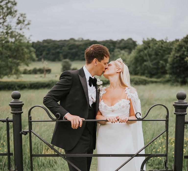 Bride in handmade wedding dress and bridal cape with large crown kisses the groom in black tie for their outdoor wedding 