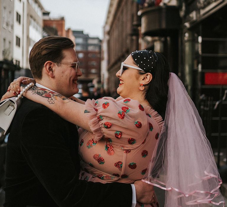 Bride wears pink veil and pink chiffon dress with glitter strawberries as her groom picks her up on their wedding day