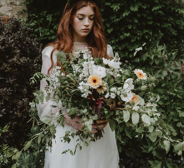 Bride in a sheer long sleeve wedding dress holding an oversized green foliage, white and yellow flower wedding bouquet 