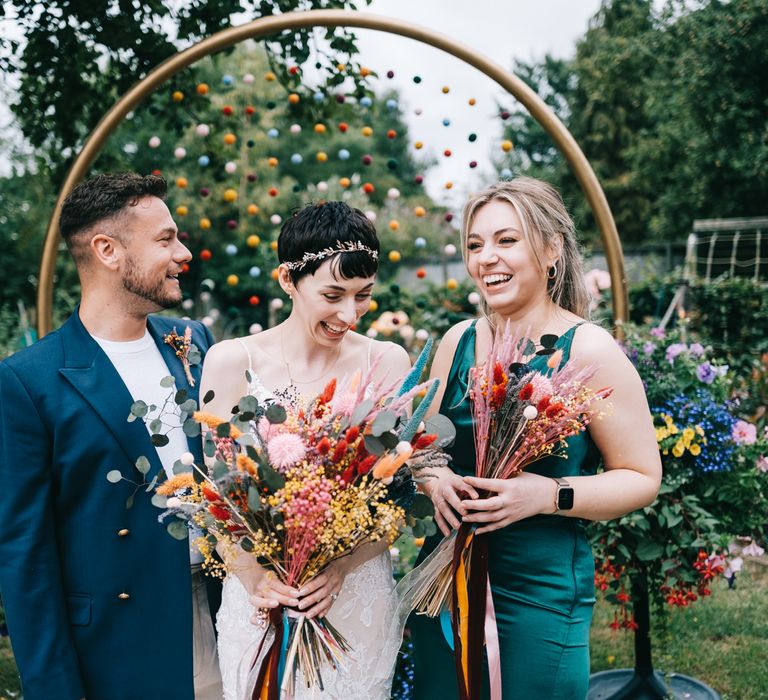 Bride holds bright bridal bouquet and stands with friends in front of DIY pom pom decorations wedding arch 