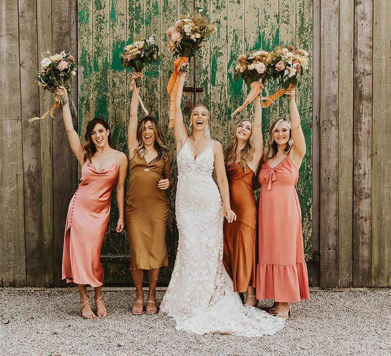 Bride in lace Made with Love wedding dress and bridesmaids in mismatched dresses lifting their bouquets in the air 