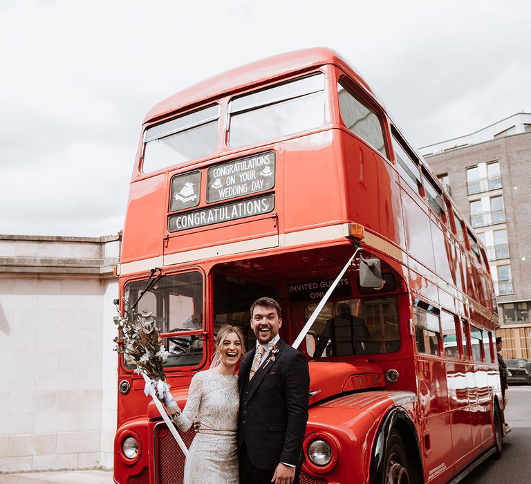 Bride in long sleeve lace wedding dress stands with her groom in front of London red bus with white ribbon to the front