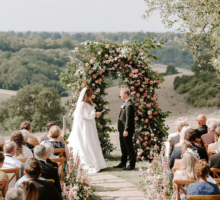 Bride & groom stand in front of beautiful floral archway with pops of orange during outdoor wedding ceremony