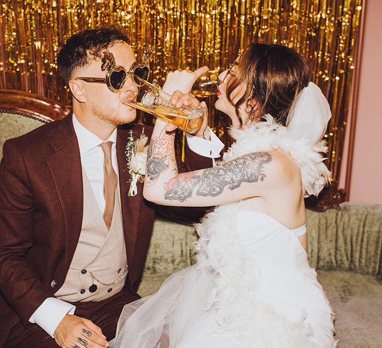 Bride & groom sit in DIY photo booth wearing fun sunglasses and gold streamers to the back