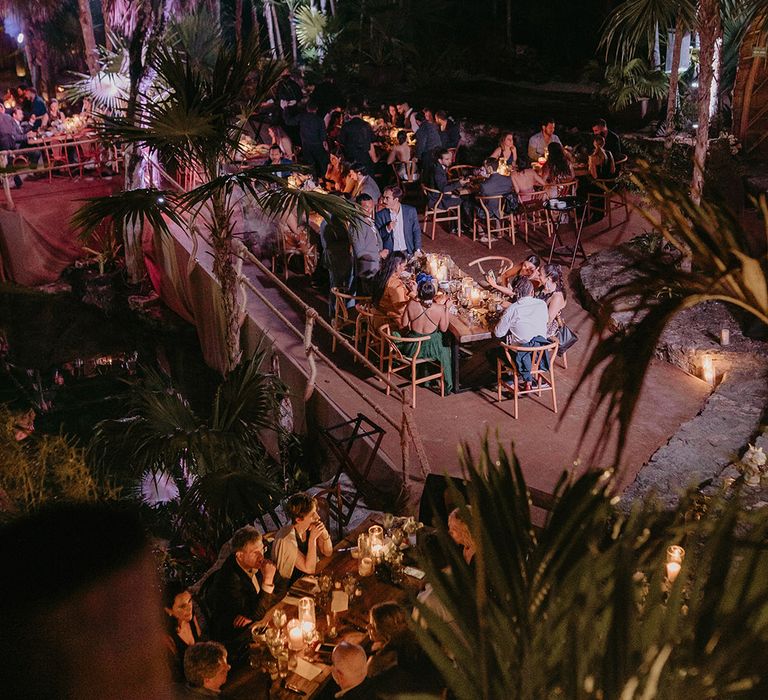 Wedding guests sit around wooden banquet tables in cenote wedding Tulum