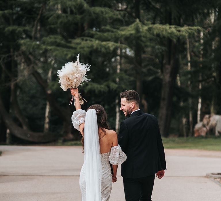 Bride in boho lace wedding dress with train raises her white dried flower bouquet as she walks away with the groom in a black suit 