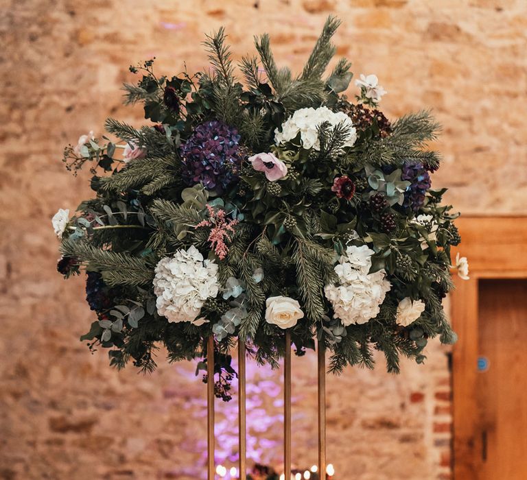 Pink anemone, white and purple hydrangea, fir tree sprigs and foliage make up a tall flower arrangement with burgundy coloured glassware and taper candles