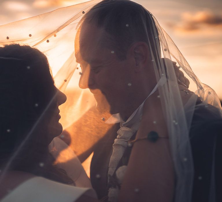 Sunset wedding photography with the bride wearing a pearl veil draped over her and the groom for a close up moment 