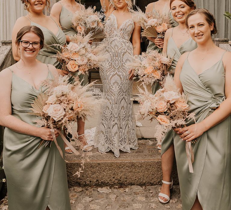 Bride in a boho lace fitted Willowby by Watters wedding dress with the bridal party in sage green satin bridesmaid dresses all holding pink rose and pampas grass bouquets 