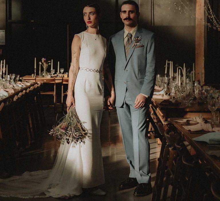 Lisa in fitted pre-loved wedding dress with open back holding DIY floral bouquet and standing beside Gabriel in powder blue Gucci suit 