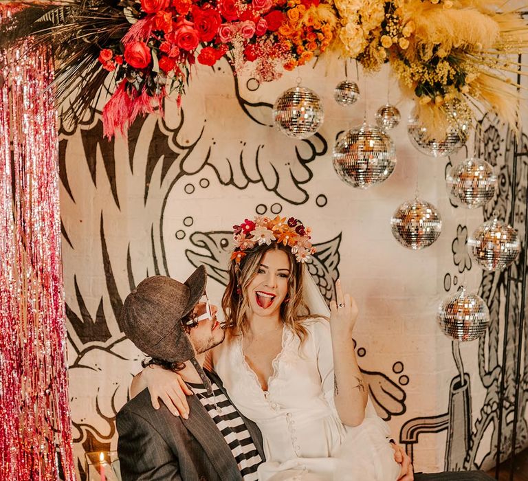 Groom in dark grey pinstripe blazer and black and white long sleeve t shirt, dark grey cap and white sunglasses with bride in three quarter length sheer sleeve wedding dress with button and cinched waist details and pink bridal flower crown and open-toe fluffy pink wedding heels sitting underneath disco balls and pink and yellow rose floral arrangements 