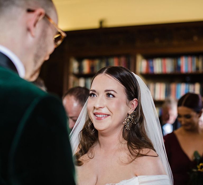 Bride gets emotional and smiles at the groom who wears a velvet green suit jacket as they stand at the alar together 