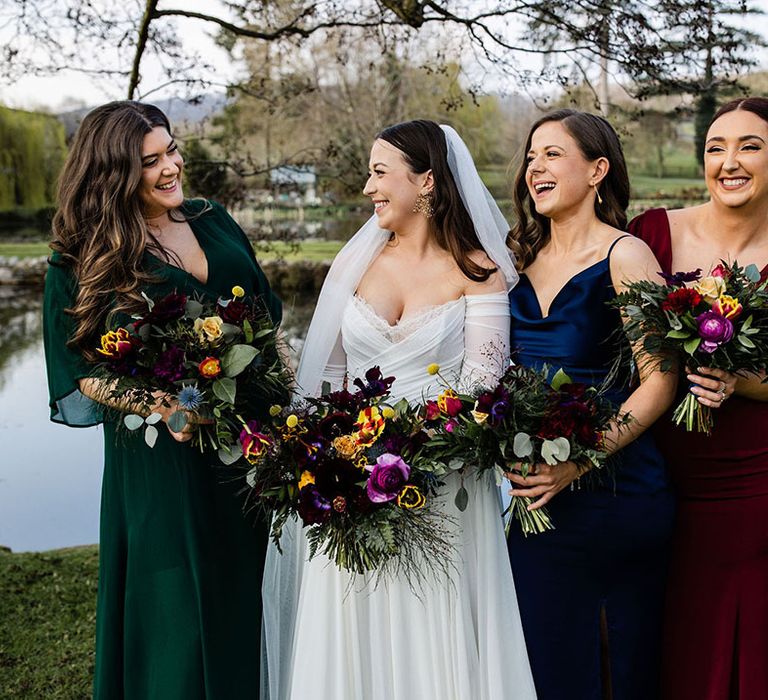 The bridal party with the bridesmaids in jewel tone mismatched bridesmaid dresses laughing with the bride holding purse and yellow spring wedding bouquets 