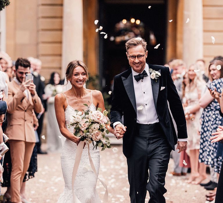 The bride wears a fitted lace wedding dress with the groom in a black tuxedo with a white rose buttonhole walking for their confetti exit from the church ceremony 