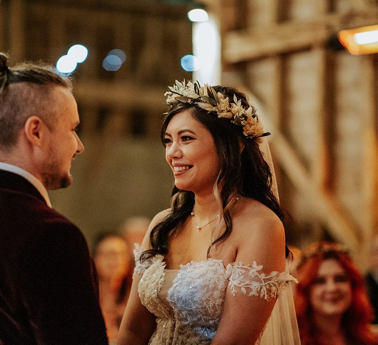 The bride in a delicate lace off the shoulder wedding dress with boho dried flower crown stares into the groom's eyes for their ceremony