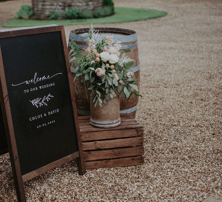 Black chalkboard welcome wedding sign with wooden crate and vintage milk churn with baby pink and white roses with greenery and dried grass with wooden barrel for rustic wedding