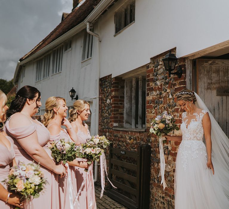 Bride in illusion lace wedding dress with her blonde hair in braided hairstyle with bridesmaids in pink bridesmaid dresses seeing her for the first time 