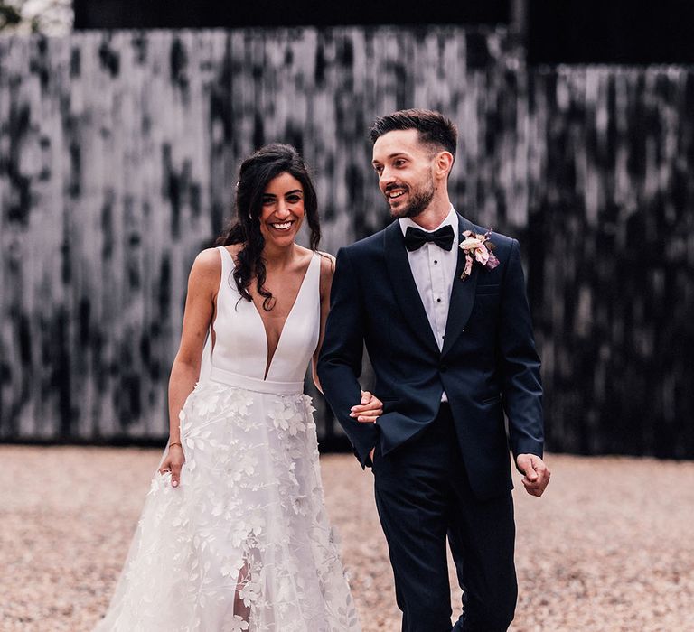 Bride in floral appliqué front slit wedding dress walking along with the groom in traditional black tie for their luxe barn wedding 